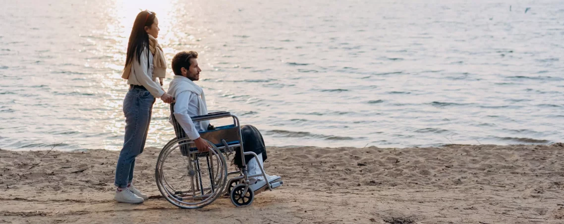 Bild Rollstuhl: Berufsunfähigkeitsversicherung Beratung - Mann sitzt im Rollstuhl und wird von Frau über den Strand geschoben
